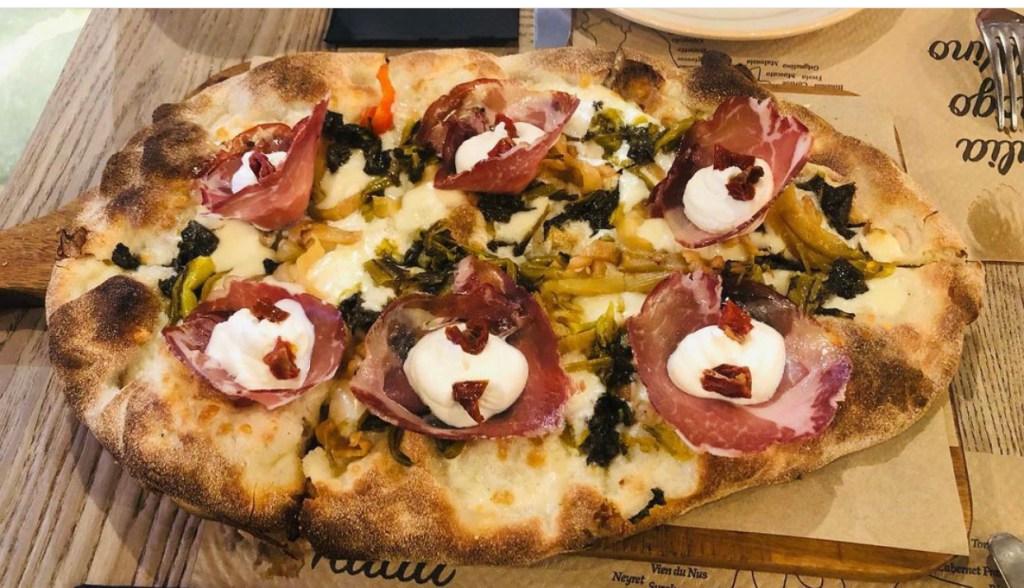 Pinsa Coppa compuesta por embutido copa, mozzarella, provola ahumada, queso burrata, tomatitos secos, grelos y berenjena encurtidas. Gastrobar Gustazio